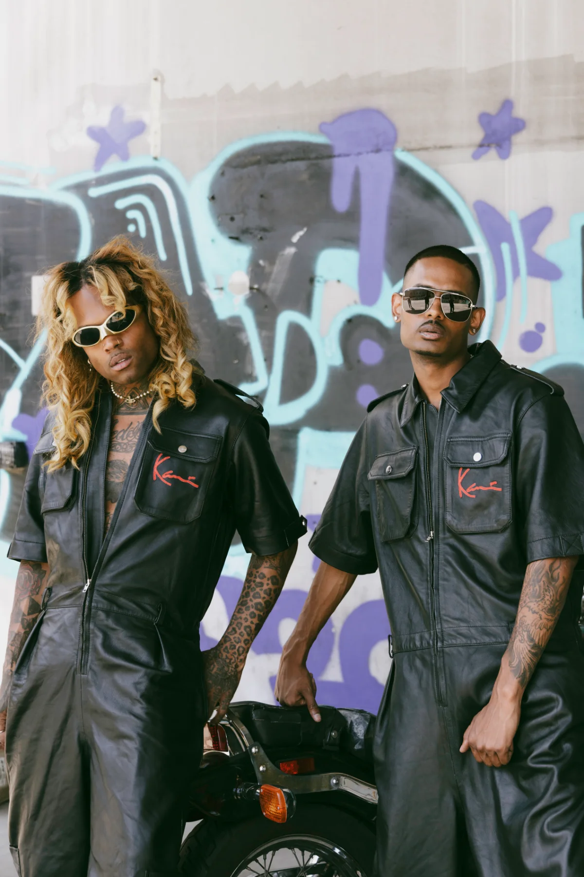 Two-up in matching black Karl Kani KK 2 Racing leather coveralls, motorcycle and graffiti wall